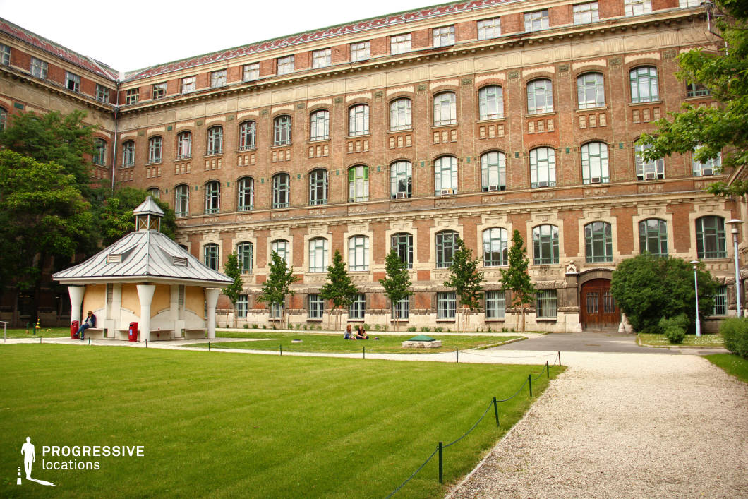 This university courtyard features a grand historic brick building with neoclassical detailing. The facade is adorned with symmetrically arranged large arched windows, decorative stone elements, and green-framed glass panes that add to the academic and institutional character. The well-maintained lawn and gravel pathways create a tranquil setting, complemented by young trees and neatly trimmed bushes. A small pavilion with a distinctive metal roof and curved supports introduces a contrasting architectural style within the courtyard. The overall atmosphere is serene and scholarly, making it an ideal location for academic, historical, or intellectual-themed settings.