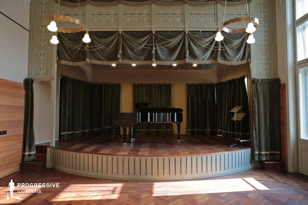 This intimate chamber music stage features a semicircular wooden platform with herringbone flooring, framed by deep green velvet curtains that add a sense of sophistication. The ornate ceiling detailing, decorative drapery, and vintage-style pendant lights enhance the space’s historical elegance. A grand piano sits at the center, accompanied by a music stand and chairs, emphasizing the room’s purpose for classical performances or refined gatherings. The warm natural light filtering through tall windows further highlights the rich textures and inviting ambiance, making this an ideal venue for recitals, lectures, or cultural events in a distinguished setting.