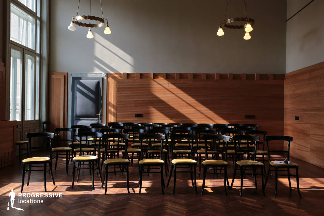 This sophisticated chamber music room features classic black wooden chairs arranged in rows on a rich herringbone parquet floor, creating an intimate yet formal atmosphere. The warm wood-paneled walls contrast with soft gray tones, while large windows allow natural light to cast striking shadows across the space. Vintage-inspired pendant lights add to the refined ambiance, complementing the high ceilings and decorative molding. The combination of historical elegance and acoustic-friendly design makes this room ideal for small concerts, lectures, or cultural events requiring an intimate and distinguished setting.
