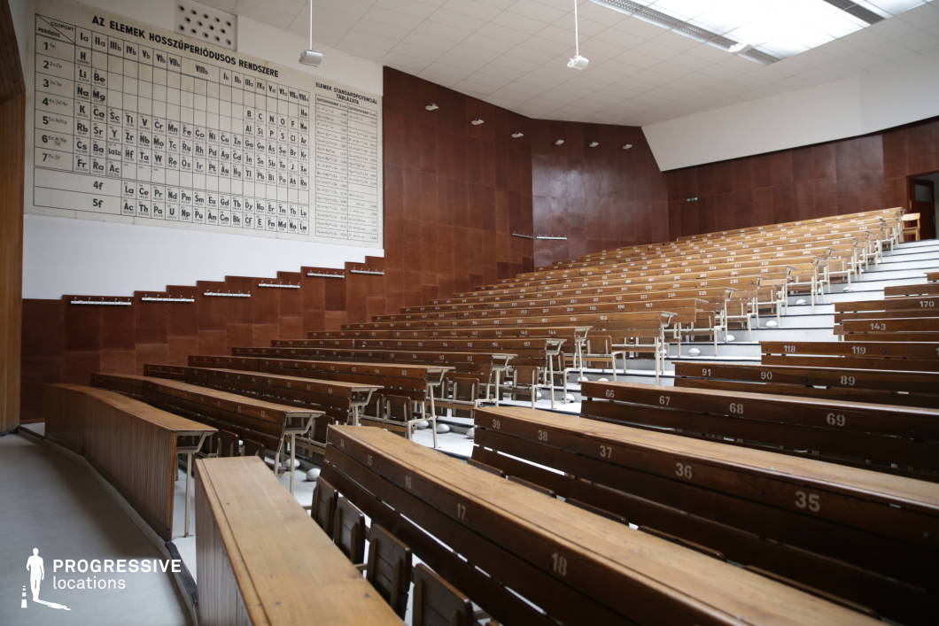 This traditional chemistry lecture hall features tiered wooden seating with numbered desks, creating a structured and academic atmosphere. The large periodic table on the front wall reinforces its educational purpose, while the rich wooden paneling on the walls adds warmth and a sense of history. A high ceiling with a skylight provides natural illumination, complementing the artificial lighting fixtures. The hall’s functional yet vintage aesthetic makes it an ideal setting for scientific lectures, academic presentations, or historically themed educational scenes.