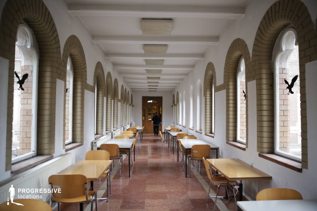This elegant university corridor features a series of symmetrical arched windows framed with light brick, allowing natural light to filter through and enhance the warm-toned stone flooring. The long, narrow layout is lined with simple wooden tables and chairs, creating an inviting study or dining space. White walls and exposed ceiling beams add to the historic yet minimalist aesthetic, while glass doors at the far end provide a focal point that extends the perspective. The combination of traditional architecture and functional furnishings makes this corridor ideal for academic settings, quiet discussions, or reflective study environments.