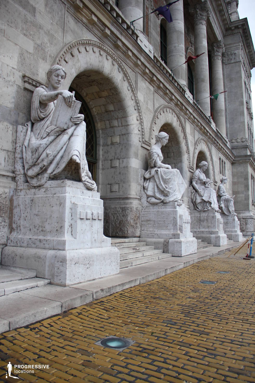 This grand neoclassical entrance features a series of intricately carved marble statues seated on large pedestals, each depicting allegorical figures in flowing robes. The imposing stone facade is characterized by arched windows, fluted columns, and decorative moldings, reinforcing the building’s historical and academic prestige. The cobblestone pavement with embedded lighting elements adds texture and depth, while the mounted flags introduce a dynamic visual contrast. The combination of classical sculptures and monumental architecture makes this location ideal for settings requiring institutional grandeur, cultural significance, or historical depth.