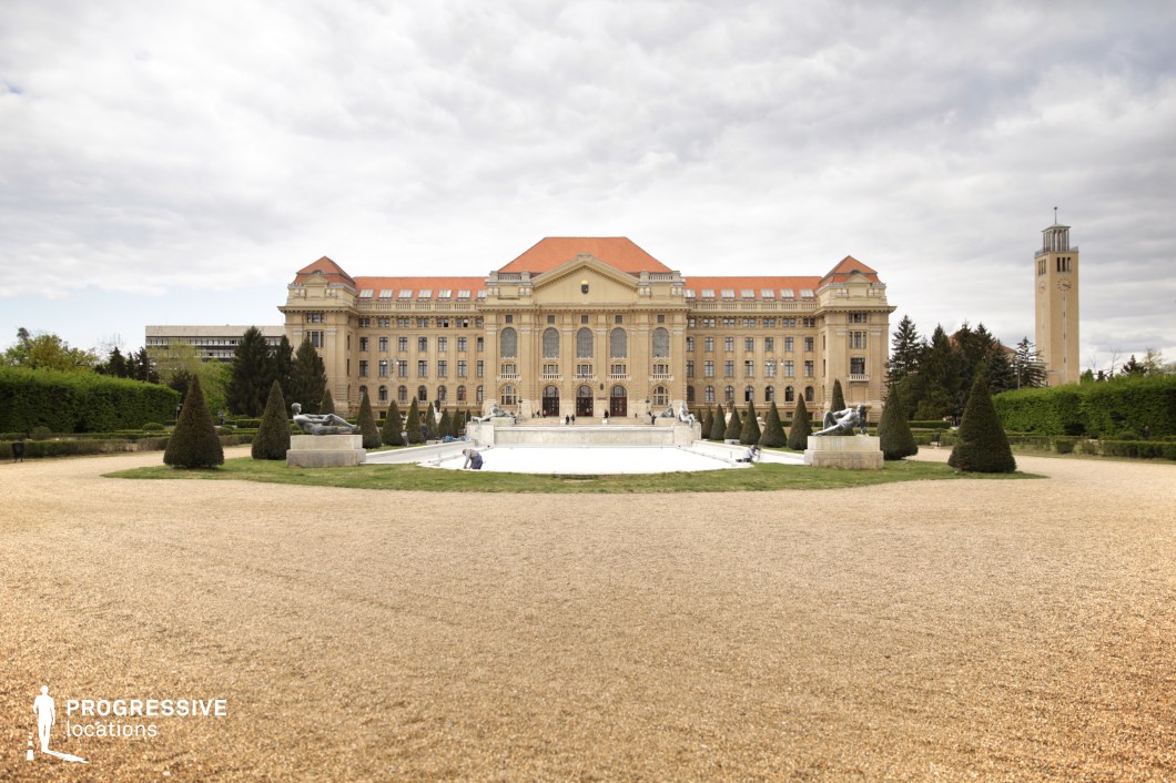 A stately neoclassical building dominates the scene, characterized by symmetrical architecture, tall arched windows, and a red-tiled roof. The facade is adorned with intricate detailing, evoking a prestigious and institutional atmosphere. In the foreground, a well-manicured formal garden with gravel pathways, sculpted shrubs, and decorative fountains adds to the grandeur. The centerpiece is a large, tiered fountain with sculptural elements, flanked by symmetrically arranged conical trees. To the right, a slender clock tower with minimalist design contrasts with the elaborate main structure. The open courtyard and balanced layout make this location ideal for scenes requiring a historical, academic, or governmental backdrop.