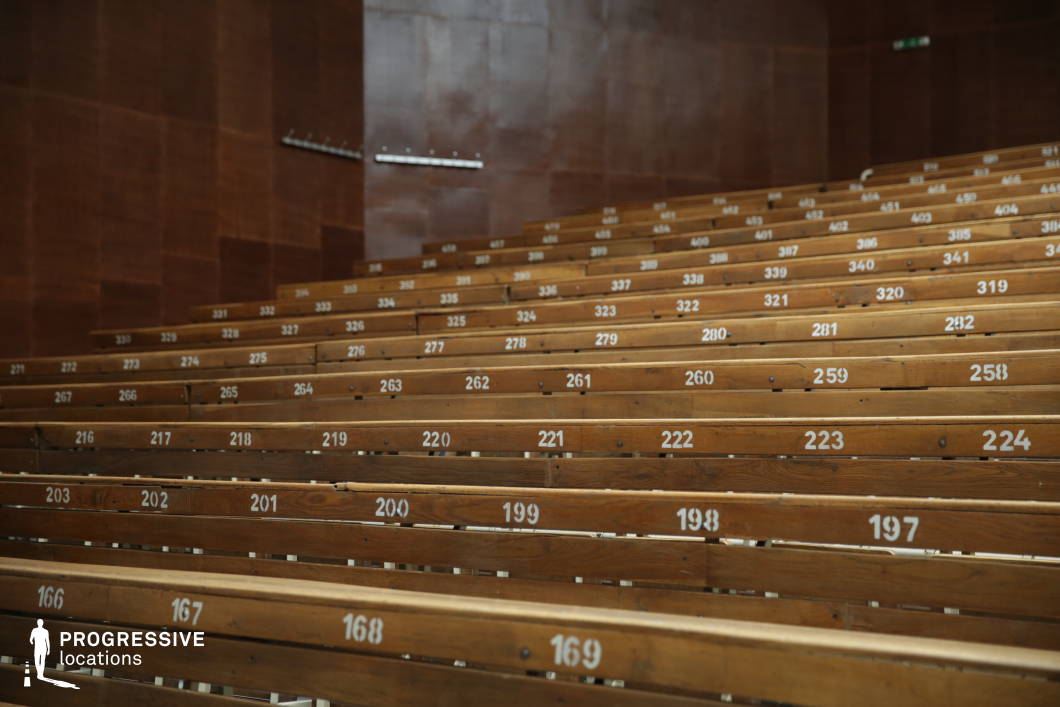 This classic lecture hall features tiered wooden bench seating with numbered placements, creating a structured and academic atmosphere. The warm-toned wooden paneling on the walls enhances the historic character, while the worn surfaces add a sense of time and tradition. The high, linear arrangement of seats ensures clear sightlines, making the space functional for large lectures or presentations. The minimal yet immersive design makes this setting ideal for educational institutions, historic academic environments, or retro-inspired cinematic scenes.