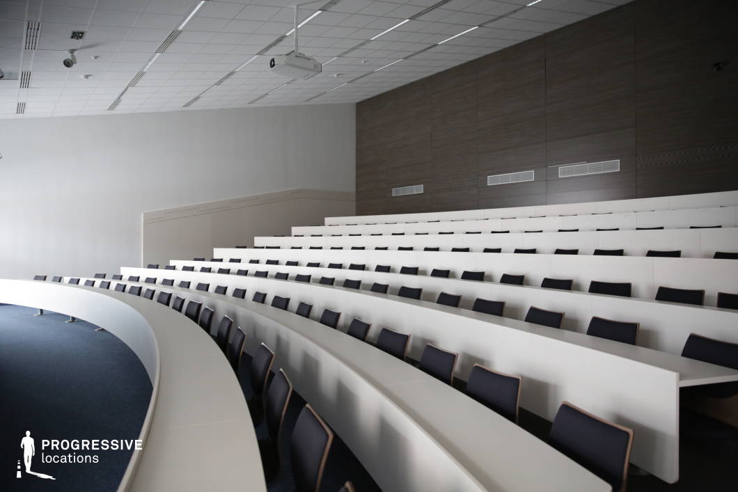 This sleek, modern lecture hall features tiered curved seating with crisp white desks and black cushioned chairs, creating a clean and professional aesthetic. The neutral color palette is complemented by dark wood-paneled walls, adding warmth and sophistication. A ceiling-mounted projector ensures high-tech presentation capabilities, while recessed lighting maintains a well-balanced ambiance. The curved design enhances visibility and engagement, making this space ideal for academic lectures, business conferences, or formal presentations in a contemporary setting.