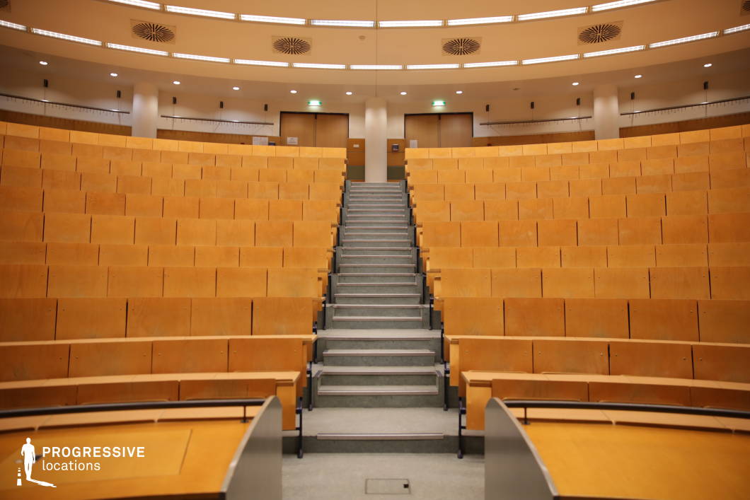 This contemporary lecture hall features a circular, tiered seating arrangement designed for optimal visibility and engagement. The pale wooden seating is neatly organized in rows, facing a central focal point at the front of the room. A staircase in the center leads to the elevated sections, providing easy access to the higher seats. The clean lines and minimalistic design of the space, with its modern lighting fixtures and unobtrusive columns, create a professional and focused atmosphere. Ideal for educational, corporate, or conference settings, this lecture hall exudes a modern, functional elegance suited for both large and small events.