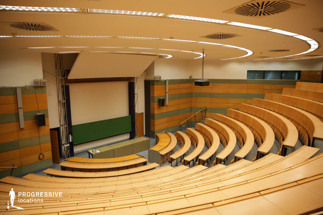 This modern lecture hall features curved, tiered wooden seating that creates a dynamic and immersive learning environment. The walls incorporate warm wood paneling with green acoustic elements, enhancing both aesthetics and sound quality. A large projection screen and a traditional chalkboard provide versatility for presentations, while recessed ceiling lighting and a suspended projector ensure optimal visibility. The curved architectural design and well-organized layout make this space ideal for academic lectures, conferences, or professional events requiring a contemporary yet functional setting.