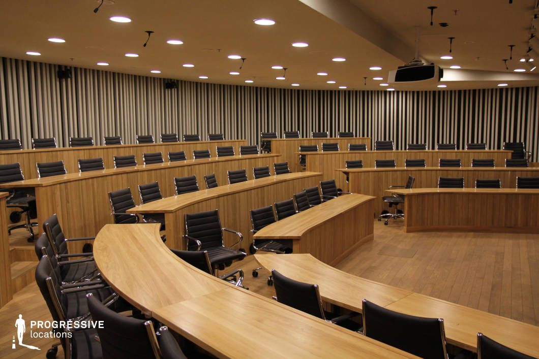 This contemporary auditorium features a tiered semicircular seating arrangement with sleek wooden desks and black leather chairs, creating a professional and collaborative atmosphere. The striped acoustic wall panels enhance both aesthetics and sound quality, while recessed ceiling lighting ensures even illumination. A ceiling-mounted projector and strategically placed microphones provide high-tech presentation capabilities. The combination of warm wood tones, modern furnishings, and an efficient layout makes this space ideal for executive meetings, academic seminars, or corporate training sessions requiring a sophisticated and functional setting.