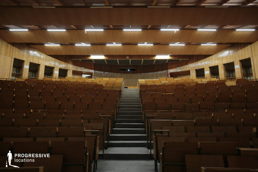 This symmetrical auditorium features tiered wooden seating with integrated foldable desks, creating a structured and immersive academic setting. The curved wooden-paneled walls and ceiling enhance the acoustics while contributing to a warm, mid-century aesthetic. A central aisle divides the seating, leading toward a stage area framed by vertical slatted wood paneling that adds depth and texture. Recessed lighting fixtures provide even illumination, making this venue well-suited for academic lectures, conferences, or formal presentations requiring a classic yet functional environment.