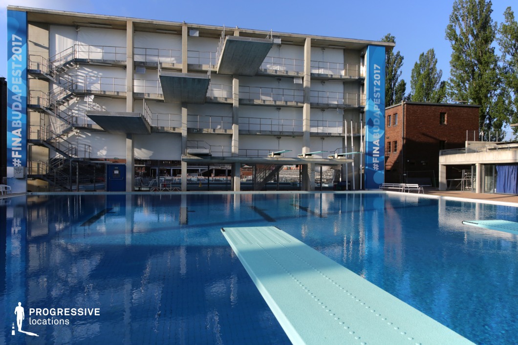 A high-performance diving pool featuring multiple diving platforms and springboards set against a modernist concrete structure. The facility has an industrial aesthetic, with metal staircases and railings providing access to the elevated platforms. The clear blue water reflects the surrounding architecture, enhancing the dramatic depth of the scene. Large banners indicate its use for international competitions, making it a premier setting for professional diving events. The open-air environment, with surrounding trees and natural light, balances the raw, functional structure. Ideal for sports-related productions, professional training visuals, or high-intensity competition scenes.