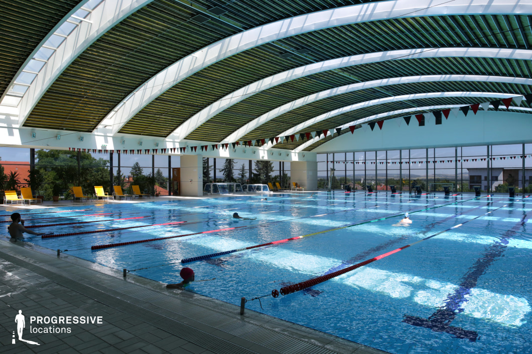 This contemporary indoor swimming pool features a striking arched ceiling with a combination of wooden and white structural beams, creating a dynamic yet elegant design. Large floor-to-ceiling windows flood the space with natural light while offering panoramic views of the surrounding landscape. The spacious pool is equipped with lane dividers, making it suitable for both leisure and professional training. Yellow lounge chairs and potted plants enhance the relaxing atmosphere. This setting is ideal for filming high-end sports training scenes, wellness retreats, or sleek modern architecture-focused productions.