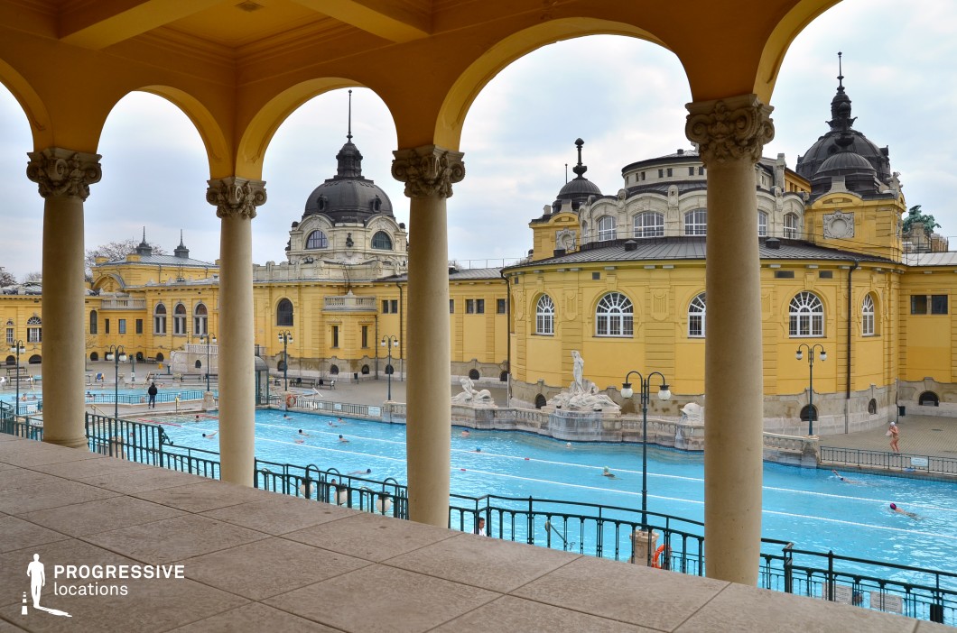 This stunning thermal bath complex showcases an opulent neoclassical architectural style with vibrant yellow facades, intricate domes, and arched windows. The foreground is framed by a series of elegant columns with ornate capitals, adding depth and symmetry to the composition. The expansive outdoor pool area is lined with decorative sculptures and wrought-iron railings, enhancing the historical grandeur of the setting. The location exudes a timeless European charm, making it ideal for filming period dramas, historical documentaries, or sophisticated travel and luxury-themed productions.