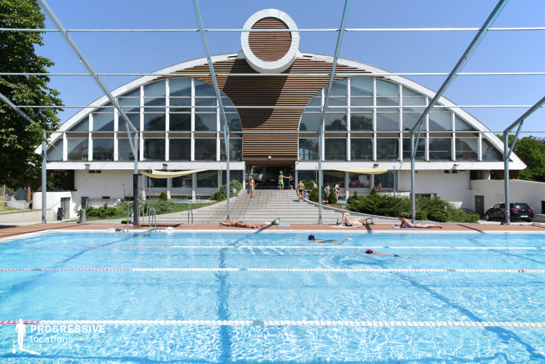 This contemporary swimming complex features a striking arched building with expansive glass windows and wooden panel accents, creating a sleek yet inviting architectural design. The symmetrical facade is complemented by a wide entrance staircase leading directly to the pool deck. The Olympic-sized outdoor pool, divided into lanes, is surrounded by a spacious wooden deck where visitors relax under the sun. A partially constructed or retractable frame structure adds versatility, suggesting seasonal adaptability. The setting is ideal for filming modern sports facilities, leisure scenes, or upscale fitness and wellness productions.