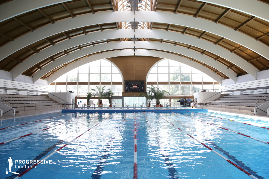 A spacious indoor swimming facility featuring a striking arched wooden ceiling with white beams, creating a visually dynamic structure. The large floor-to-ceiling windows allow abundant natural light to flood the space, enhancing the reflections on the clear blue water. The pool is marked for lanes, making it ideal for competitive swimming or training. Surrounding the pool, tiered seating provides a spectator-friendly environment. Tropical plants near the pool deck add a welcoming touch. This location is well-suited for sports-related productions, wellness themes, or high-end architectural cinematography.