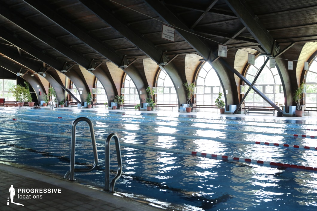 This unique indoor swimming pool features a series of curved wooden arches that create a warm and inviting atmosphere. Large arched windows line the walls, allowing natural light to filter in and reflect off the water’s surface. The dark timber ceiling contrasts with the bright outdoor views, adding depth to the space. Potted plants along the poolside enhance the tranquil and natural ambiance. This location is ideal for filming serene training montages, period dramas, or wellness-themed productions, offering a visually rich and architecturally distinctive setting.
