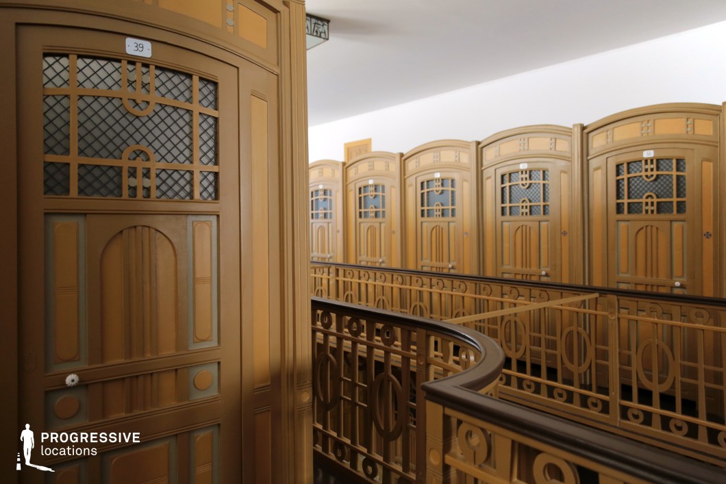 A stunning arrangement of vintage wooden dressing cabins, showcasing Art Nouveau-inspired craftsmanship. The space is defined by intricately designed cabin doors featuring geometric and curved motifs, framed by warm brown tones with subtle green and gold accents. Each door incorporates decorative glass panels with a lattice design, adding to the refined aesthetic. A curved wooden railing with matching ornamental details guides along the upper level, emphasizing the architectural elegance of the space. The softly illuminated interior and harmonious symmetry make this an ideal setting for period film productions, high-end fashion editorials, or historical documentary visuals.