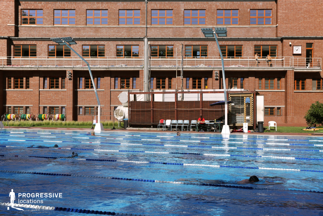 This professional-grade outdoor swimming pool is set against a striking brick building with large windows and metal railings, creating an industrial yet functional sports facility aesthetic. The pool is divided into multiple lanes with floating markers, while modern overhead lighting structures enhance the competition-ready environment. A shaded seating area and a row of training equipment along the edge suggest a high-performance training atmosphere. This location is ideal for filming sports documentaries, training montages, and competitive swimming or water sports-themed productions.