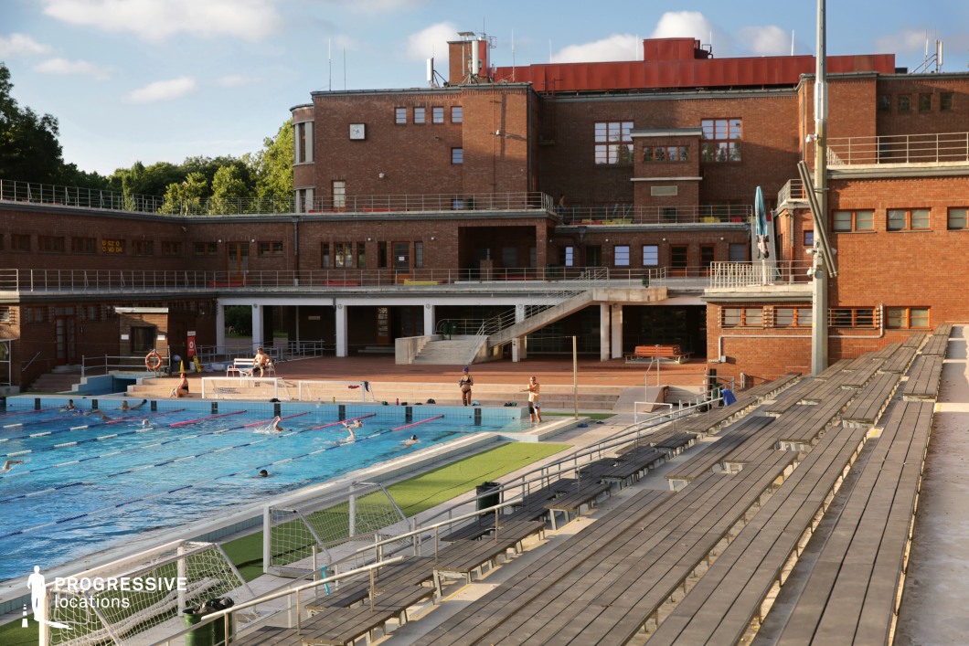 This expansive outdoor swimming complex features a large Olympic-style pool set against a bold, industrial-style brick building with metal railings and functional staircases. The stadium-style wooden bleachers provide ample seating, reinforcing the competitive sports atmosphere. The structure’s geometric lines and warm brick tones contrast with the cool blue of the water, creating a visually dynamic setting. This location is ideal for filming sports dramas, training montages, and competitive swimming or water polo-themed productions.