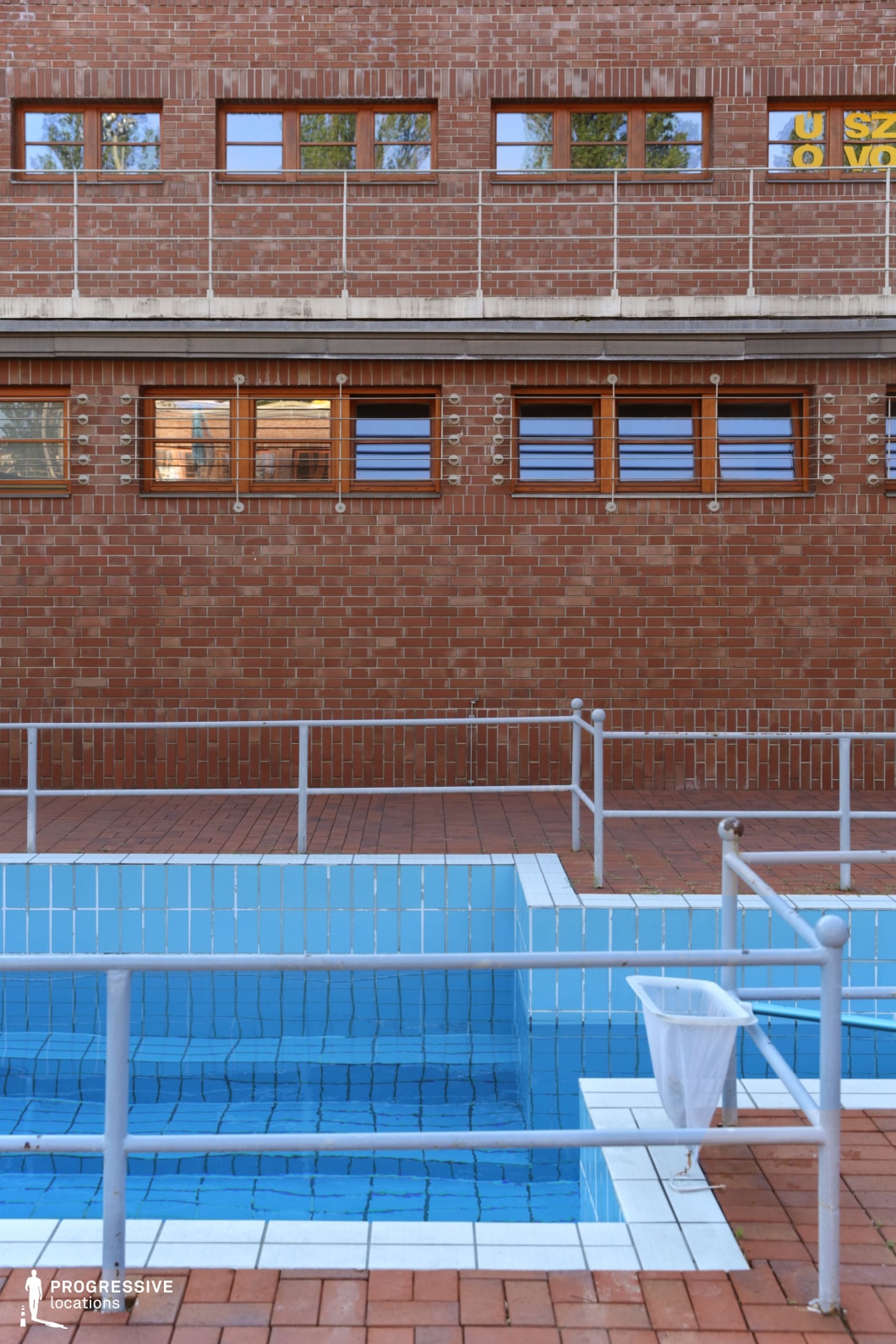 A compact outdoor swimming pool set against a striking red brick wall, featuring warm wooden-framed windows and metal railing details. The pool area is surrounded by terracotta paving, which contrasts with the cool blue tiles lining the water. A safety railing encloses the space, adding to the structured and functional design. The symmetrical arrangement of windows and architectural elements provides a balanced, industrial yet inviting atmosphere, making it an ideal setting for urban recreational scenes or architectural-focused projects.