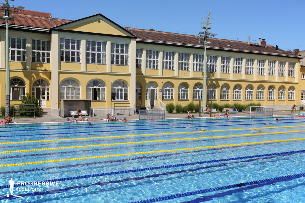 A historic outdoor swimming pool adjacent to a classical European-style building with a symmetrical yellow façade and arched windows. The architecture reflects early 20th-century design, featuring a tiled roof, large multi-pane windows, and decorative archways. The pool is divided into multiple lanes with bright yellow and blue floating dividers, emphasizing its function as a professional or recreational swimming facility. Surrounding the area, lounge chairs and benches provide a relaxed atmosphere, while tall lampposts suggest evening usability. This location is ideal for sports-focused productions, period film settings, or luxury lifestyle visuals, blending athletic functionality with a vintage architectural backdrop.