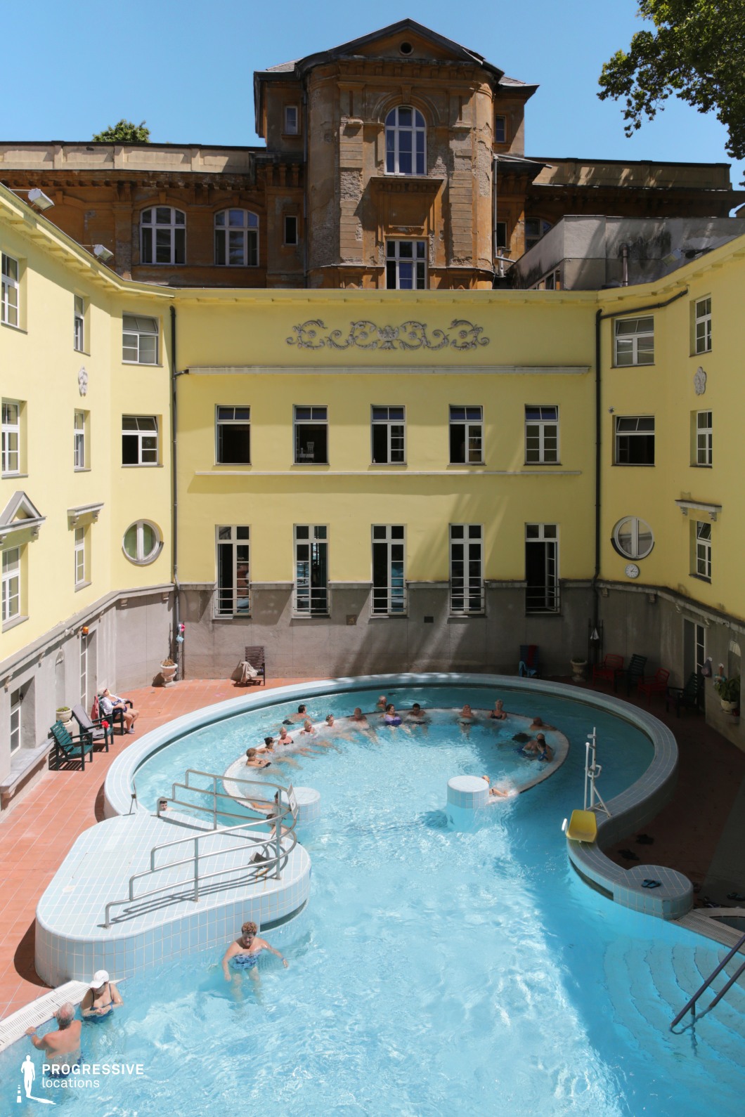 A charming outdoor thermal pool set within a courtyard enclosed by a historic yellow building. The structure exhibits classical European architectural elements, including ornate decorative reliefs and symmetrically arranged windows. In contrast, an aged stone façade in the background suggests a deeper historical significance. The pool itself has a curved design with a built-in bench for relaxation, surrounded by terracotta flooring and scattered seating areas. This location offers a rich blend of classical elegance and historic ambiance, making it a versatile setting for period-piece cinematography, wellness-focused productions, or travel and luxury lifestyle visuals.