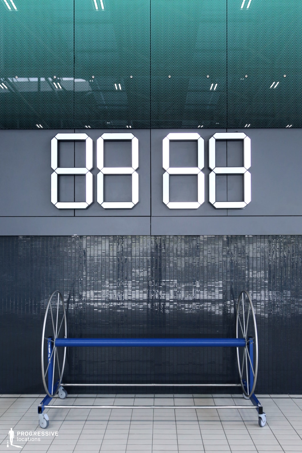 A sleek, minimalist digital clock display mounted on a tiled wall inside an aquatic sports facility. The oversized, segmented LED numbers create a bold, futuristic aesthetic against the dark reflective tiles. The upper portion of the wall features a textured green panel with a subtle wave pattern, enhancing the modern design. Below the clock, a blue and silver rolling equipment reel adds a functional element to the scene. The clean lines, high-tech ambiance, and neutral tones make this an ideal setting for competitive sports, contemporary architecture, or high-performance training visuals.