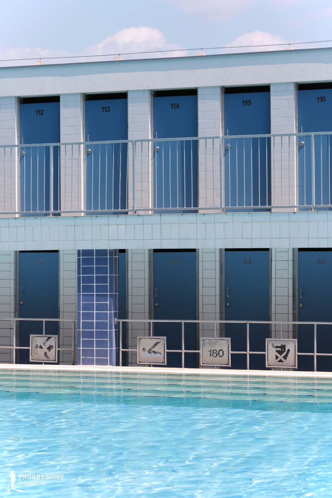 A vibrant outdoor swimming pool is framed by a two-level row of blue dressing cabins, creating a structured and functional atmosphere. The uniform doors, numbered and aligned, add to the geometric symmetry of the scene. The light blue tiled exterior complements the crystal-clear water, enhancing the fresh and inviting aesthetic. A safety railing with instructional signs borders the pool area, reinforcing the practical design. Ideal for scenes requiring a clean, minimalist backdrop with a hint of retro functionality.