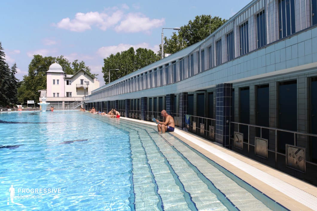A long, elegant outdoor pool is lined with blue-tiled dressing cabins, evoking a traditional bathhouse aesthetic. The stepped entry into the water creates a smooth transition, ideal for leisure and relaxation. The reflective surface of the pool mirrors the bright sky, enhancing the sense of openness. A classic white villa with a domed rooftop stands in the background, adding a historical and sophisticated touch. Metal railings with vintage-style signs provide functional guidance, reinforcing the location's structured design. This setting is perfect for scenes emphasizing relaxation, tradition, and architectural charm.