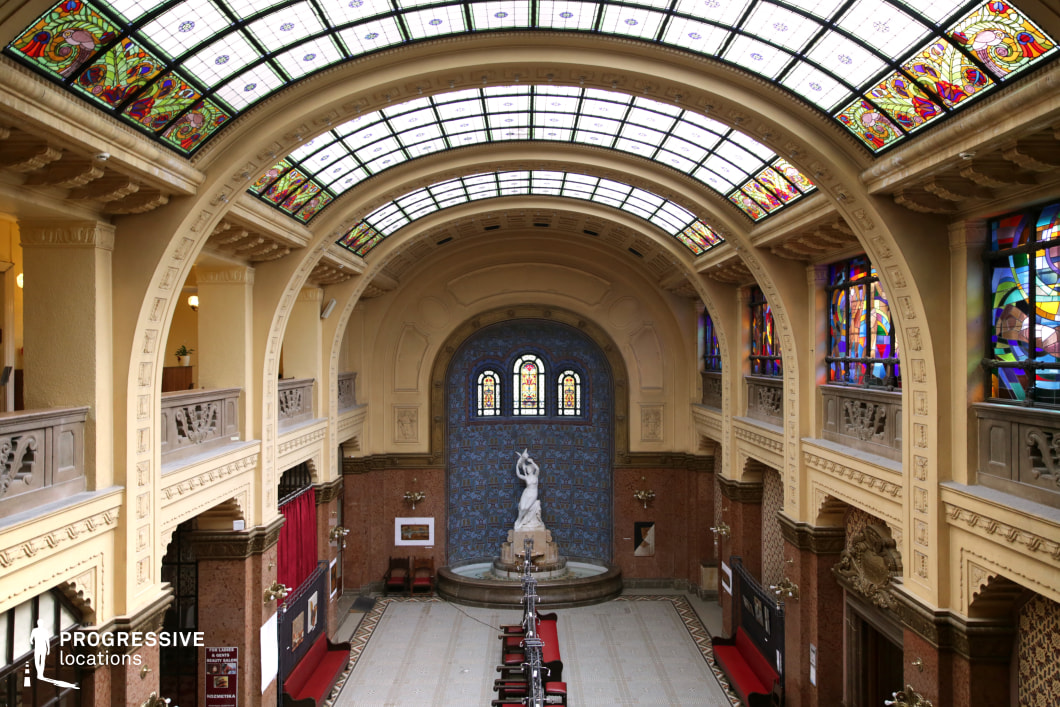 This breathtaking atrium showcases a majestic Art Nouveau design, featuring an expansive arched glass ceiling adorned with intricate stained-glass patterns that bathe the space in soft, colorful light. The high walls are lined with elegant balconies, sculpted balustrades, and decorative moldings, adding to the grandeur of the setting. At the far end, a striking statue is framed by an ornate blue mosaic wall, enhanced by arched stained-glass windows. The symmetrical composition and intricate detailing make this an ideal filming location for historical dramas, period pieces, and visually rich artistic productions.