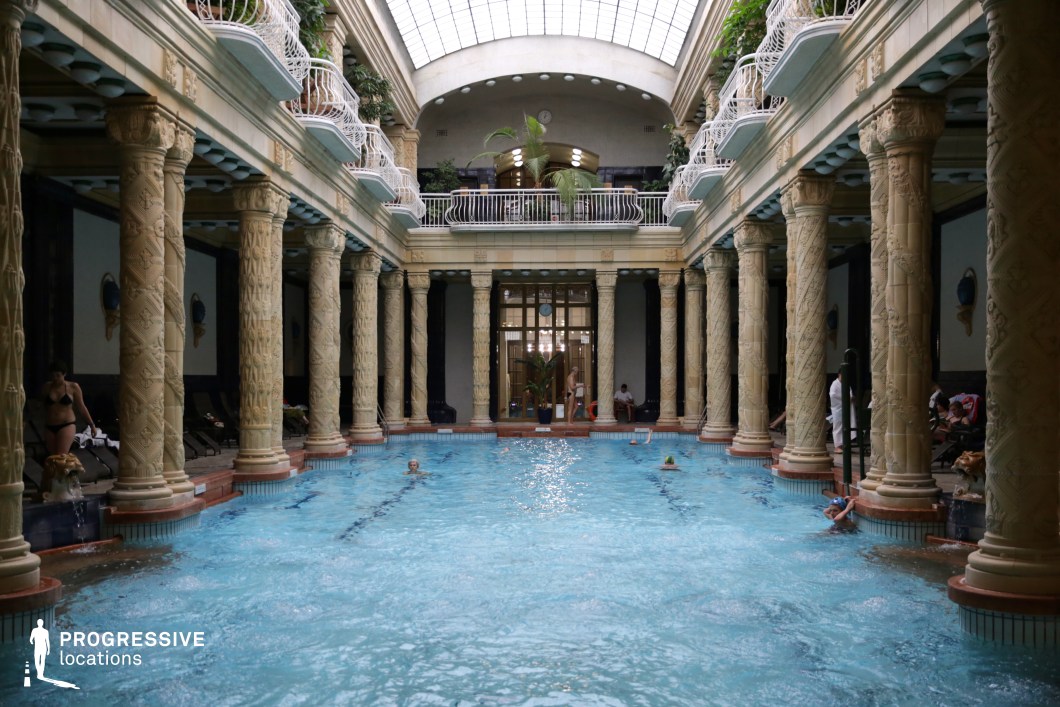 This opulent indoor pool hall features a grand classical design, highlighted by intricately carved stone columns and an expansive arched glass ceiling that floods the space with natural light. The two-story layout includes elegant white wrought-iron balconies adorned with lush greenery, enhancing the luxurious ambiance. Decorative wall sconces and sculpted lion head fountains add historical richness to the setting. The symmetrical composition and regal atmosphere make this location ideal for filming period dramas, historical documentaries, and high-end luxury or wellness productions.