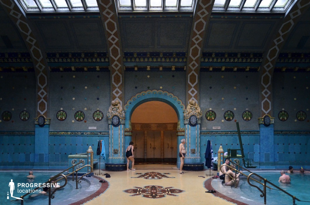 This lavish thermal bath interior features intricate Art Nouveau detailing, with rich blue and gold mosaic tiles covering the walls and floors. The grand arched doorway, adorned with sculpted lion motifs and decorative glass inlays, serves as a striking focal point. The high ceiling, supported by geometric-patterned beams, allows natural light to filter through the skylights, casting a soft glow on the space. Small circular stained-glass windows add to the historic charm, while the symmetrical design enhances the luxurious ambiance. This location is perfect for filming period dramas, historical documentaries, or fantasy productions that require an elegant and immersive setting.