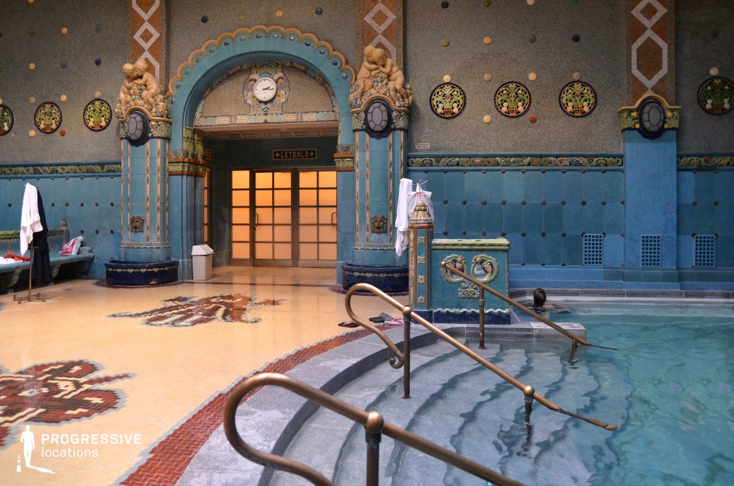 This exquisite thermal bath interior showcases intricate Art Nouveau design elements, with rich blue mosaic tiles, decorative wall patterns, and sculpted figures adorning the grand arched entrance. The symmetrical composition is emphasized by circular stained-glass accents and gold-inlaid motifs, creating a visually striking contrast against the deep blue walls. The warm-toned mosaic floor features elegant geometric patterns, while brass handrails and ornate pillars enhance the luxurious atmosphere. The combination of historical elegance and intricate detailing makes this location ideal for period dramas, historical documentaries, and visually rich cinematic productions.