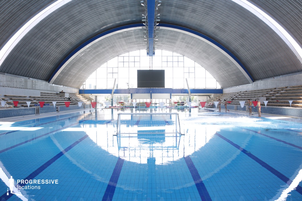 A vast indoor Olympic-sized pool designed for water polo and competitive swimming, housed within a dramatic arched metal ceiling. The reflective pool surface captures the symmetry of the curved structure, while large floor-to-ceiling windows allow natural light to flood the space. The tiered spectator seating adds a competitive sports atmosphere, complemented by banners and goal nets set for water polo. This venue is ideal for sports-related productions, professional training scenes, or dynamic aquatic cinematography requiring a sleek, industrial-modern aesthetic.