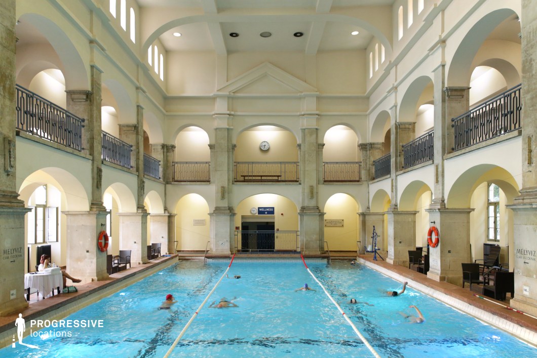 This grand indoor swimming hall features a symmetrical, neoclassical design with high vaulted ceilings, arched openings, and two levels of decorative iron railings. The cream-colored walls and stone columns create a refined and timeless aesthetic, complemented by natural light filtering through the upper windows. The elongated pool, surrounded by seating areas, adds a sense of luxury and spaciousness. The harmonious blend of classical architecture and functional design makes this an ideal filming location for historical dramas, sports-themed productions, and high-end wellness or luxury lifestyle content.