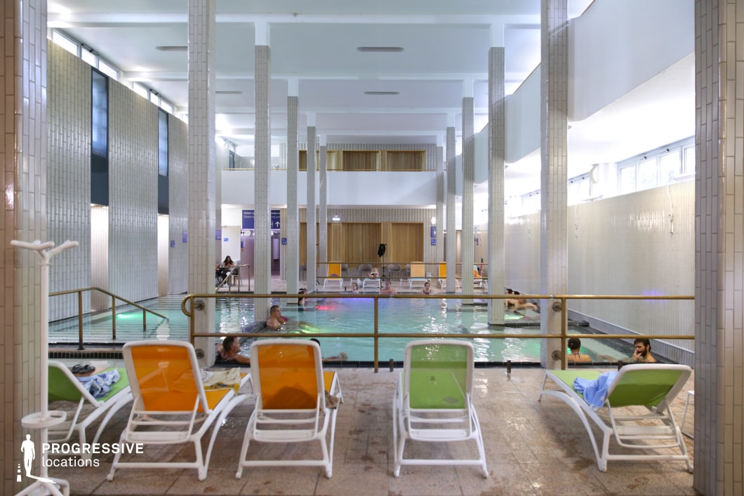 A contemporary indoor thermal pool area featuring a striking geometric architectural design. Tall, white-tiled columns evenly spaced throughout the space create a structured and symmetrical visual appeal. The pool is surrounded by colorful lounge chairs, offering a relaxed and inviting atmosphere. The walls are clad in beige and white rectangular tiles, enhancing the clean and minimalist aesthetic. Warm wooden panel accents add a touch of sophistication to the otherwise modern setting. The pool area is illuminated by natural light filtering through the upper windows, combined with soft artificial lighting. The location is well-suited for wellness and relaxation-themed productions, offering a sleek yet tranquil ambiance.