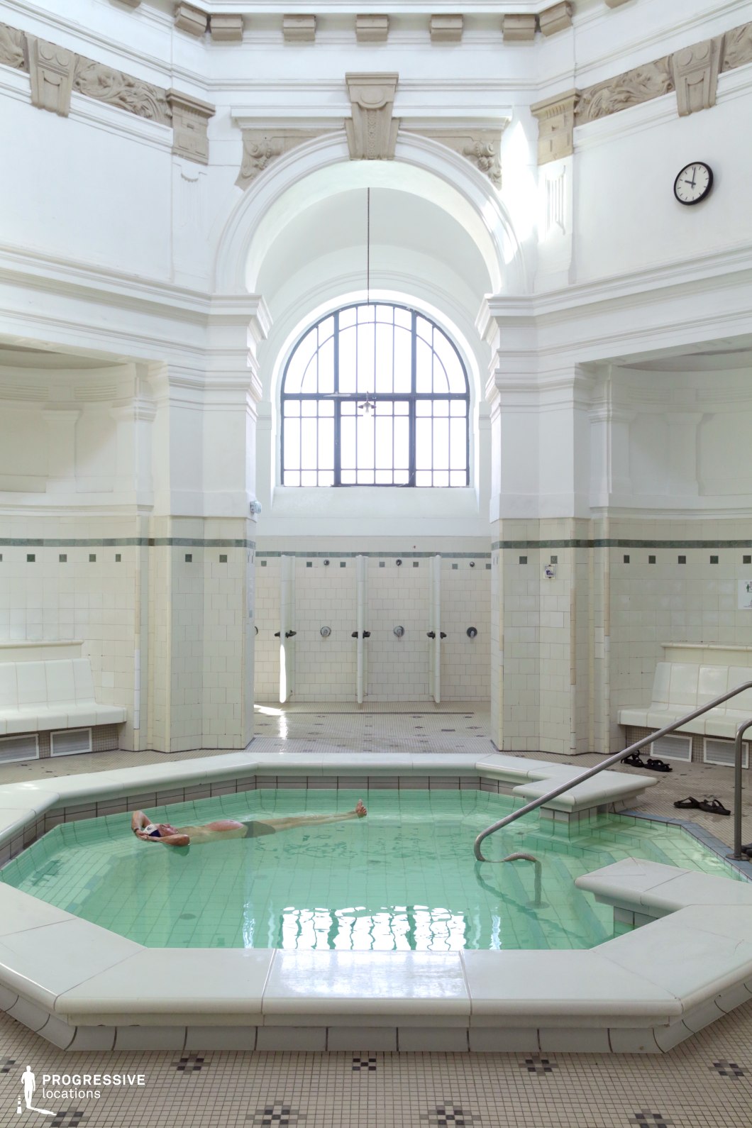 This elegant thermal bath features a serene, neoclassical design with a striking octagonal pool at its center. The high, vaulted ceiling and arched windows allow an abundance of natural light to illuminate the space, enhancing the soft white and cream-colored tiles. Ornate moldings and sculpted reliefs add historical character, while the symmetrical layout of showers and seating areas creates a sense of harmony. The bright, minimalistic aesthetic makes this location ideal for filming period dramas, wellness-focused productions, or high-end spa and luxury lifestyle content.