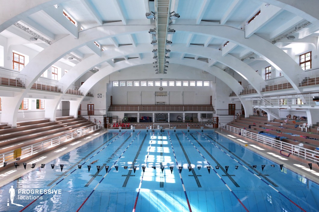 A historic indoor Olympic-sized swimming pool housed within an impressive white arched ceiling structure. The symmetrical, curved beams create a striking architectural feature, complemented by rows of tiered seating for spectators. Natural light streams in through high-set windows, enhancing the airy and spacious atmosphere. The pool is marked for competitive lanes, making it ideal for training, tournaments, or sports-related productions. A blend of classic and functional design, this location suits cinematic projects requiring a grand yet disciplined aquatic setting.