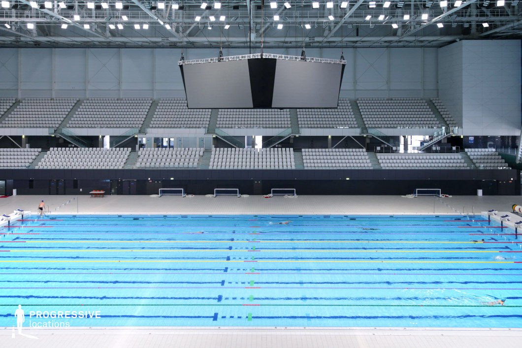 This modern Olympic swimming arena features a sleek, high-tech design with a competition-standard pool, marked lanes, and expansive tiered seating. The vast, geometric ceiling supports an array of bright overhead lights, ensuring optimal visibility for events. A suspended jumbo screen dominates the central space, reinforcing the high-profile sports atmosphere. The clean lines, monochrome seating, and state-of-the-art infrastructure make this location ideal for filming major sporting events, athlete training sequences, and high-energy competition scenes.