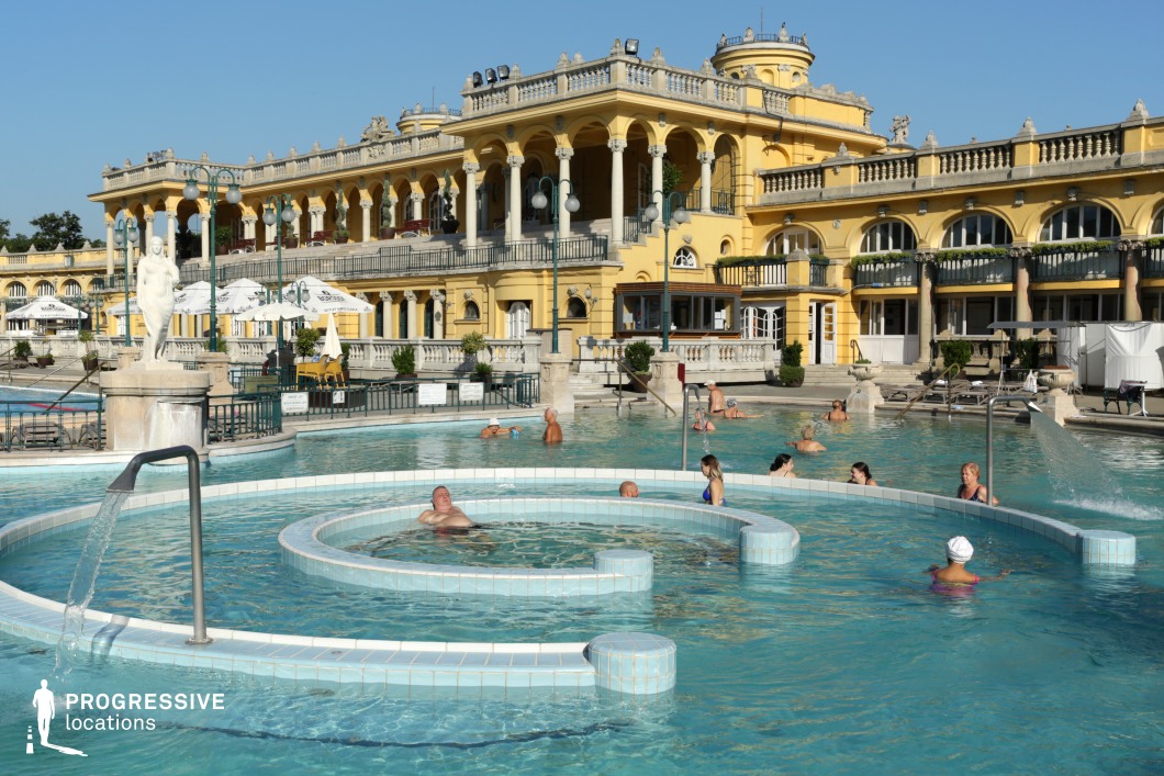 This luxurious outdoor thermal bath is framed by a grand neoclassical building with intricate balustrades, arched colonnades, and sculptural details. The pool features circular water channels and fountains, adding a sense of relaxation and sophistication. The surrounding area is enhanced with decorative lampposts, statues, and lush greenery, creating a refined European ambiance. The harmonious blend of historic architecture and water elements makes this location ideal for filming period dramas, luxury travel content, and lifestyle productions focused on wellness and leisure.