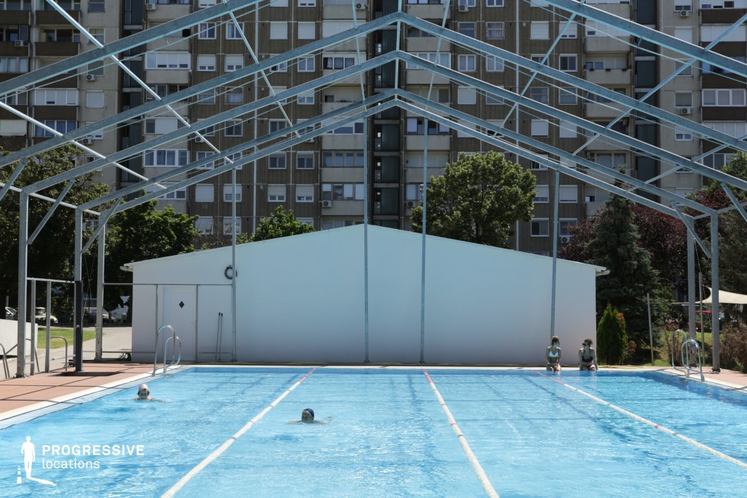 This outdoor swimming pool is set against a striking urban backdrop of a large, multi-story residential building, creating a visually compelling contrast between leisure and city life. The pool features clear blue water, divided into lanes for structured swimming. Above the pool, a partially constructed or open-frame metal structure suggests a seasonal or retractable roof system. A small white utility building at the far end adds a functional element, possibly serving as a changing area or equipment storage. Surrounding the pool, green trees and landscaped elements provide a touch of nature within the urban environment. This setting is ideal for cinematic urban recreation scenes or architectural contrasts between leisure and high-density living.