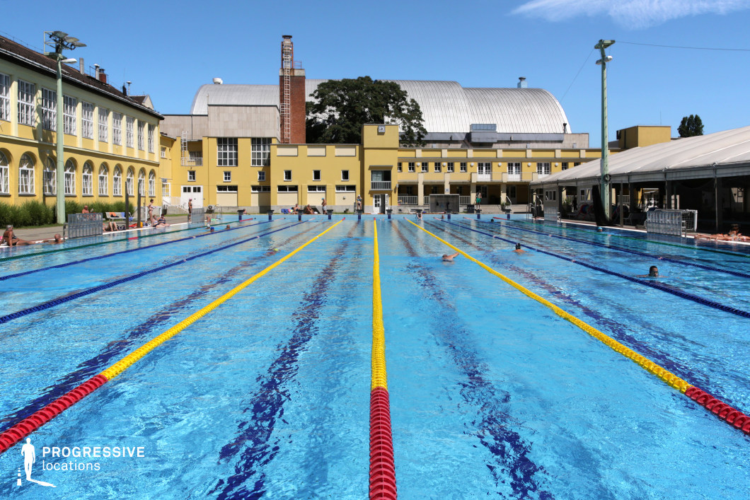 A spacious Olympic-sized outdoor swimming pool, surrounded by a mix of classical and industrial-style architecture. The vibrant blue water is divided into lanes by colorful floating markers, creating a dynamic and structured visual. On one side, a historic yellow building with arched windows adds a traditional European character, while a modern metallic-roofed structure in the background introduces an industrial contrast. The poolside area features shaded seating and lounge spaces, making this an ideal location for sports-focused productions, historical sports documentaries, or lifestyle visuals centered around elite aquatic training.
