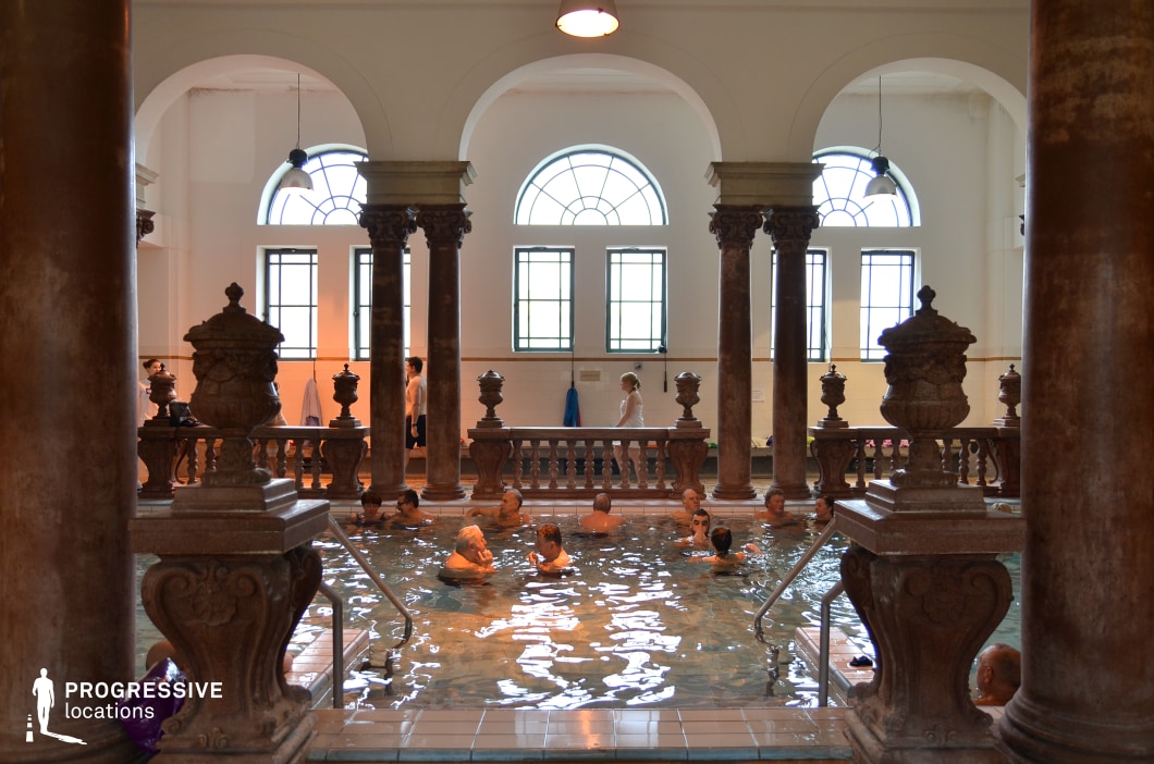 This luxurious thermal bath showcases a grand neoclassical design with towering marble columns, intricate balustrades, and arched windows that allow natural light to filter into the space. The rich brown tones of the stone contrast beautifully with the bright white walls and large windows, enhancing the refined and historic ambiance. Decorative urns and sculptural details add to the grandeur, while the water’s reflection creates a serene and immersive atmosphere. This setting is ideal for period dramas, historical documentaries, and elegant wellness or luxury-themed productions.