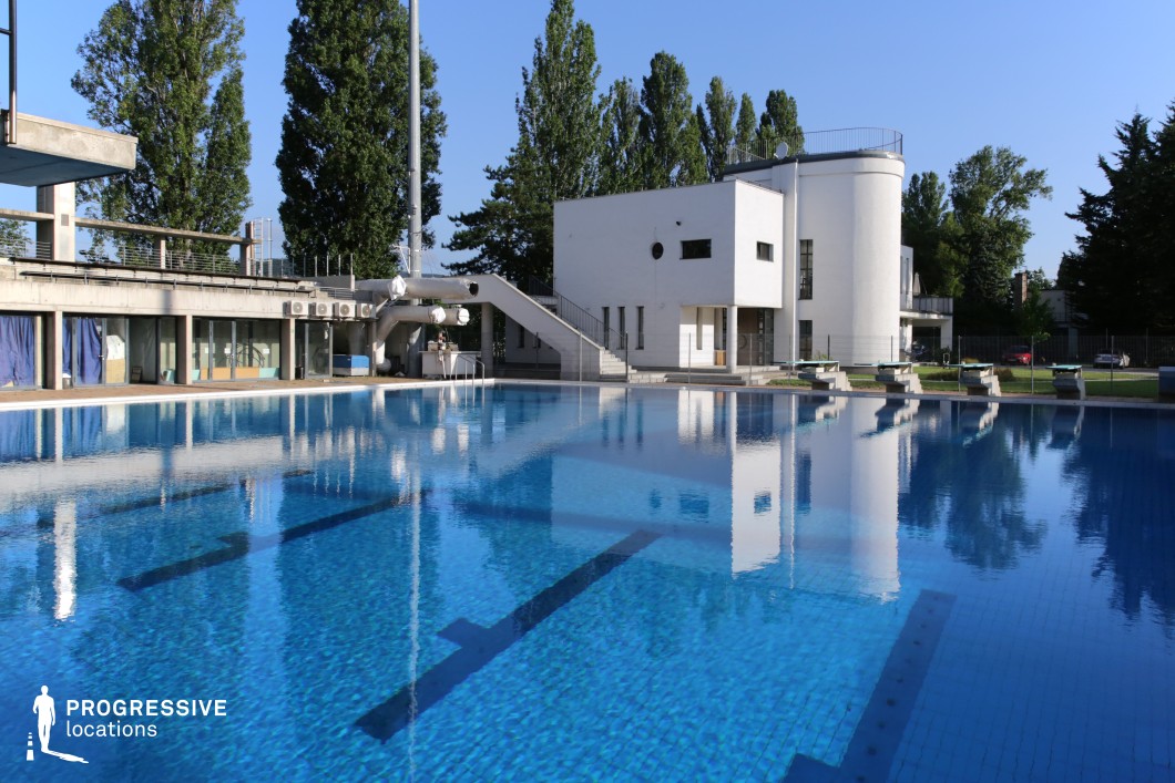 A striking outdoor swimming pool reflecting a modernist white building with clean geometric lines. The deep blue water contrasts with the bright architectural elements, enhancing the crisp, contemporary aesthetic. The pool area is equipped with diving blocks, while the surrounding greenery and tall trees provide a natural frame. A functional concrete walkway and structural elements, such as an external staircase, add an industrial touch. This setting is ideal for professional training visuals, architectural photography, or sophisticated wellness and sports productions.