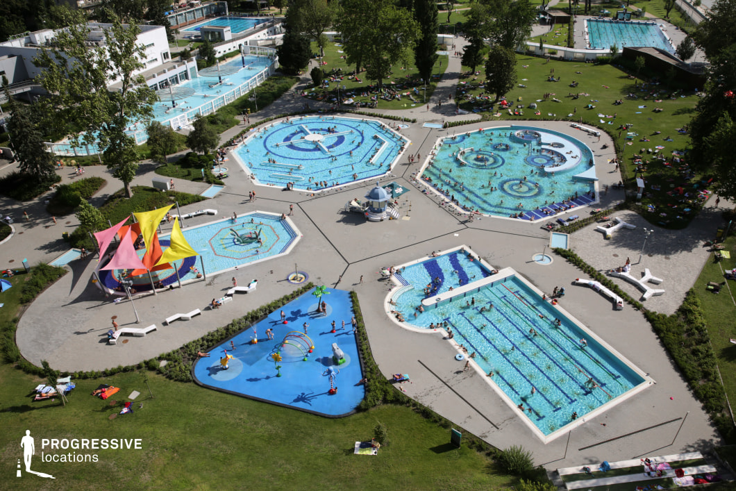 A stunning aerial perspective of a contemporary outdoor water park, showcasing a variety of geometrically shaped pools, each with unique features. The octagonal and irregularly shaped pools are adorned with intricate blue mosaic patterns, creating a visually engaging layout. A central plaza connects the pools, surrounded by green lawns where visitors relax. The area includes a children's splash zone with vibrant water features, a shaded play area with colorful fabric sails, and a dedicated lap pool with marked swimming lanes. Modern white lounge chairs and pathways contrast with the lush greenery, enhancing the park’s inviting atmosphere. This location is ideal for capturing dynamic leisure scenes, resort-style visuals, or expansive summer recreation settings.