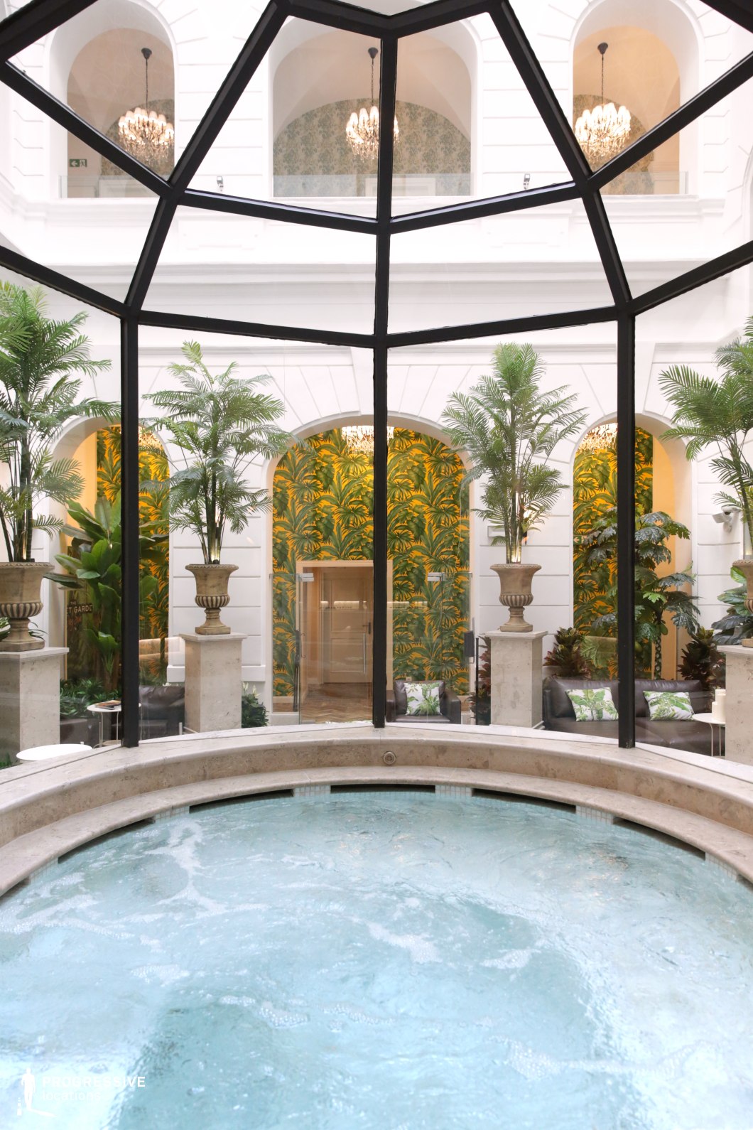 An opulent indoor spa set within a glass-enclosed atrium, blending neoclassical architecture with tropical-inspired decor. The circular jacuzzi in the foreground is encased in natural stone, its bubbling water creating a sense of relaxation. Towering potted palms and lush greenery complement the white arched walls, which feature intricate moldings and high ceilings. Ornate chandeliers cast a warm glow, enhancing the refined ambiance. The background showcases arched openings leading to a relaxation area adorned with vibrant tropical wallpaper, plush seating, and elegant stone pedestals. This spa setting exudes sophistication and tranquility, making it an ideal location for high-end wellness and hospitality productions.