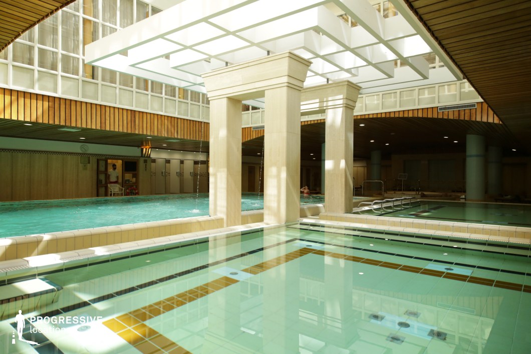 This contemporary indoor spa features a sleek architectural design with clean lines, geometric columns, and a combination of warm wood paneling and cream-colored tiles. Large windows allow natural light to filter through, enhancing the reflections on the water’s surface. The pool area is divided into sections, with cascading water features adding a dynamic visual element. The mix of open and covered spaces creates a balanced ambiance between relaxation and modern luxury. This location is ideal for filming wellness and spa-themed productions, high-end travel content, and contemporary lifestyle visuals.