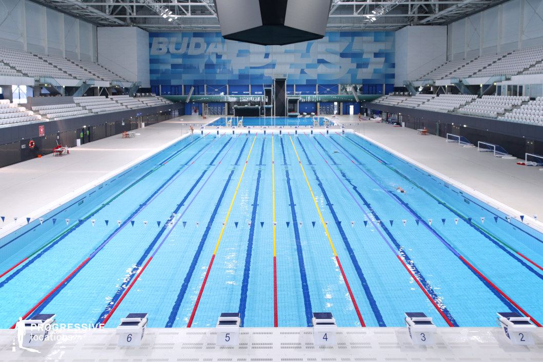 This state-of-the-art Olympic swimming arena features a pristine competition pool with clearly marked lanes, surrounded by tiered grandstands with sleek white seating. The expansive, high-ceilinged structure is illuminated by bright overhead lighting, while a large digital scoreboard and bold blue branding on the back wall emphasize the professional sports setting. The symmetrical architecture and clean, minimalist aesthetic enhance the high-performance atmosphere. This location is ideal for filming international swimming competitions, sports documentaries, and motivational training sequences.