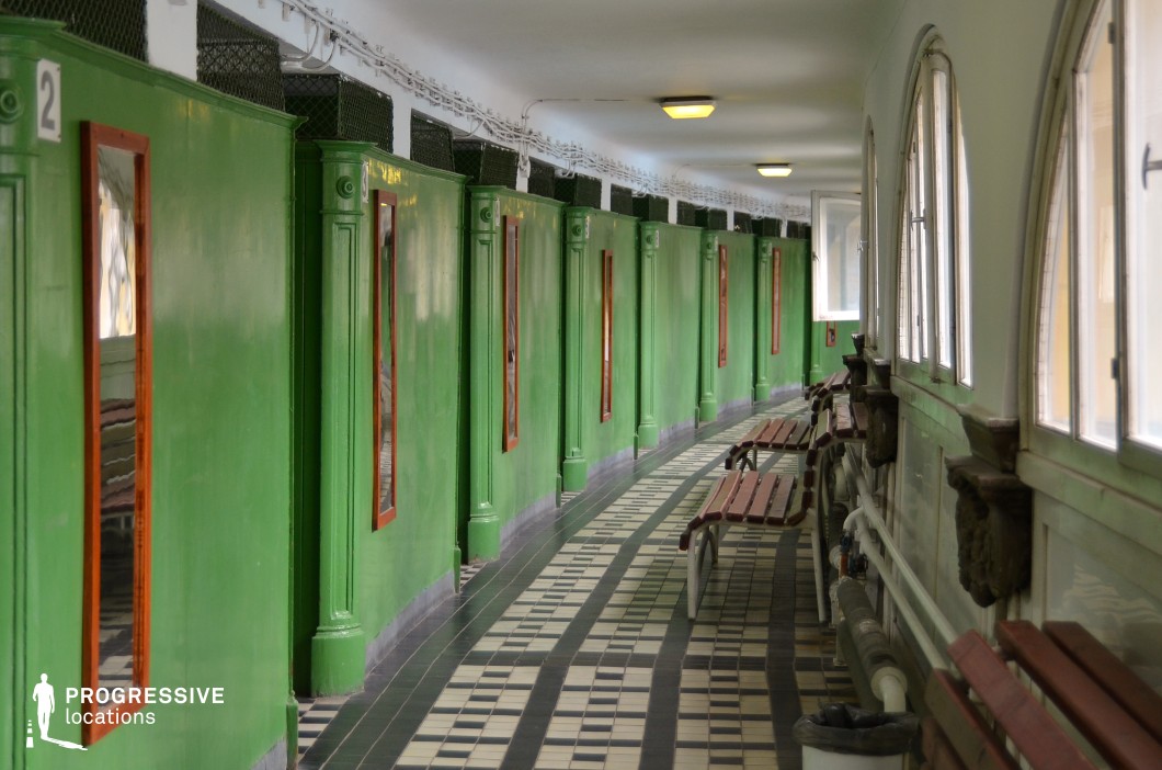 A historic dressing cabin corridor with bold green wooden partitions, each featuring a rectangular mirror framed in natural wood. The curved hallway is lined with a checkerboard-patterned tile floor in black and white, adding a strong visual rhythm. Large arched windows on one side allow natural light to filter in, while a series of aged wooden benches with metal supports create a resting area. The overhead lighting, exposed pipes, and wire-mesh ventilation above the cabins contribute to the space's old-world charm. This location is ideal for period film settings, vintage fashion photography, or architectural documentary projects.