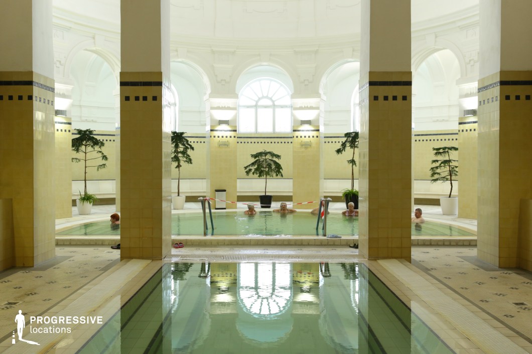 This refined indoor thermal bath features a harmonious blend of classical architecture and modern spa aesthetics. Tall, arched windows allow soft natural light to illuminate the domed ceiling, while pale yellow tiles and white decorative moldings create a serene and sophisticated ambiance. The symmetrical layout, with grand pillars dividing the pools, enhances the sense of balance and tranquility. The reflections in the still water add depth to the composition, while potted plants introduce a touch of greenery. This location is ideal for filming period dramas, high-end wellness productions, and luxurious lifestyle content.