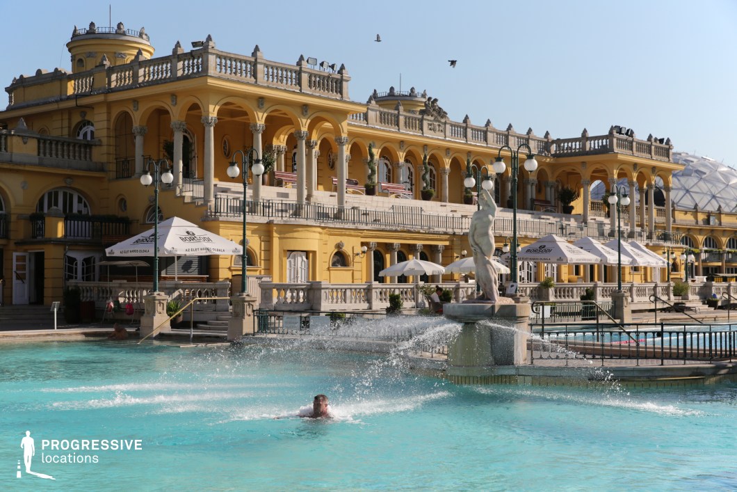 This magnificent outdoor thermal bath is set against a backdrop of an opulent Baroque-style building with vibrant yellow facades, elegant arched colonnades, and intricate balustrades. The pool area is enhanced by classical sculptures and decorative fountains that contribute to the grandeur of the setting. Ornate street lamps and shaded seating areas under umbrellas add to the refined atmosphere. The symmetry and historical elegance of the architecture make this an ideal filming location for period dramas, luxury lifestyle productions, and romantic or historical-themed films.
